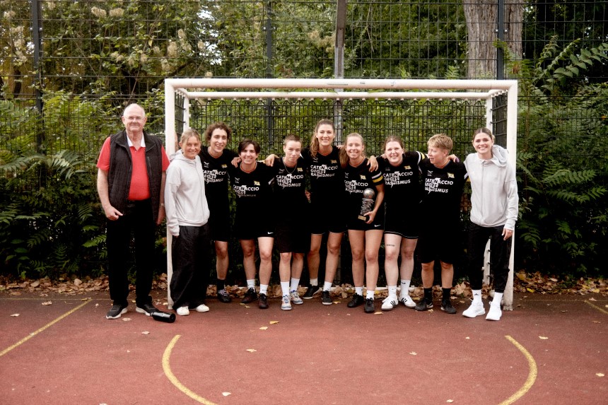 Das Foto zeigt eine Gruppe von Personen, die eng zusammen vor einem kleinen Fußballtor auf einem Outdoor-Spielfeld stehen. Alle tragen überwiegend schwarze T-Shirts mit weißem Aufdruck. Die Gruppe steht auf einem roten Bodenbelag, der typisch für einen Bolzplatz oder Street-Soccer-Platz ist. Die Personen haben ihre Arme umeinander gelegt, was auf Teamzusammenhalt hinweist. Einige tragen kurze Hosen und Sportschuhe, andere lange Hosen. Links und rechts neben der Gruppe stehen zwei Personen in anderer Kleidung: eine Person in dunkler Hose und weißem Oberteil, die andere in heller Sportkleidung mit weißen Schuhen. Im Hintergrund ist ein hoher Metallzaun zu sehen, der den Platz umgibt, dahinter dichtes grünes Gebüsch und Bäume. Auf dem Boden liegen einige Blätter, was auf eine herbstliche Jahreszeit schließen lässt. Die Szene vermittelt den Eindruck eines Mannschaftsfotos nach einem Street-Soccer-Turnier.