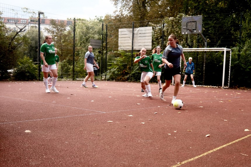 People playing soccer on a red hard pitch. In the background are trees and a goal. Photo: Lisa Loriene