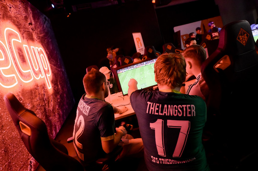 The picture shows a gaming environment with several people sitting at computers and playing a soccer video game. In the foreground, two people can be seen from behind, both wearing dark jerseys with white prints. On the left jersey is the number "12" and a name, on the right jersey the number "17" and the name. Both are sitting in large gaming chairs in front of a monitor on which a soccer pitch can be seen. The scene is bathed in warm, reddish-orange light emanating from large, glowing lettering on the left wall. It reads "eCup" in large letters. In the background, there are other people at similar gaming stations. The atmosphere is reminiscent of an e-sports tournament or gaming event.