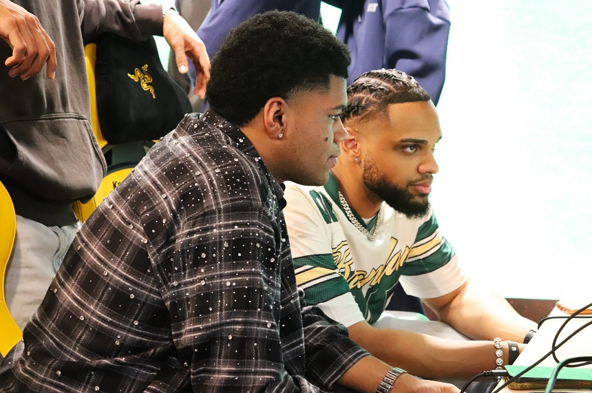 Auf dem Foto sitzen zwei Personen dicht nebeneinander an einem Tisch oder einer Konsole. Die Person links trägt ein dunkles, kariertes Oberteil mit weißen Punkten. Sie lehnt sich leicht nach vorn, als würde sie sich konzentriert einem Gespräch oder einer gemeinsamen Aktivität widmen. Die Person rechts trägt ein gestreiftes Oberteil in Grün‑ und Beigetönen mit einem grafischen Schriftzug auf der Brust sowie eine Halskette und ein Armband. Ihre Hände befinden sich vor ihr auf dem Tisch, als würde sie etwas bedienen oder steuern. Im Hintergrund sind weitere Personen unscharf zu erkennen, die stehen oder vorbeigehen. Die Szene wirkt wie ein gemeinsamer Moment in einem Gaming‑, Technik‑ oder Freizeitbereich. Die Atmosphäre vermittelt Austausch und Zusammenarbeit.