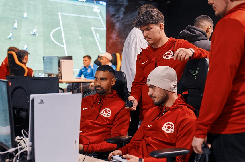 In einem Raum mit gedämpfter Beleuchtung sitzen mehrere Personen an Gaming‑Plätzen. Im Vordergrund sind drei Personen zu sehen, die rote Trainingsjacken mit einem runden Logo tragen. Zwei von ihnen sitzen auf Gaming‑Stühlen vor Monitoren und Konsolen‑Controllern, während eine dritte Person danebensteht und sich leicht nach vorn beugt, als würde sie etwas erklären oder unterstützen. Auf den Tischen stehen Monitore, Konsolen und weiteres Gaming‑Equipment. Im Hintergrund befinden sich weitere Personen, ebenfalls an Computern oder Konsolenarbeitsplätzen. Auf einer großen Leinwand oder einem Bildschirm ist eine Szene eines Fußball‑Videospiels zu erkennen, die den Gesamtraum dominiert. Die Atmosphäre wirkt konzentriert und gemeinschaftlich, als würde ein E‑Sport‑Training oder ein Turnier stattfinden.
