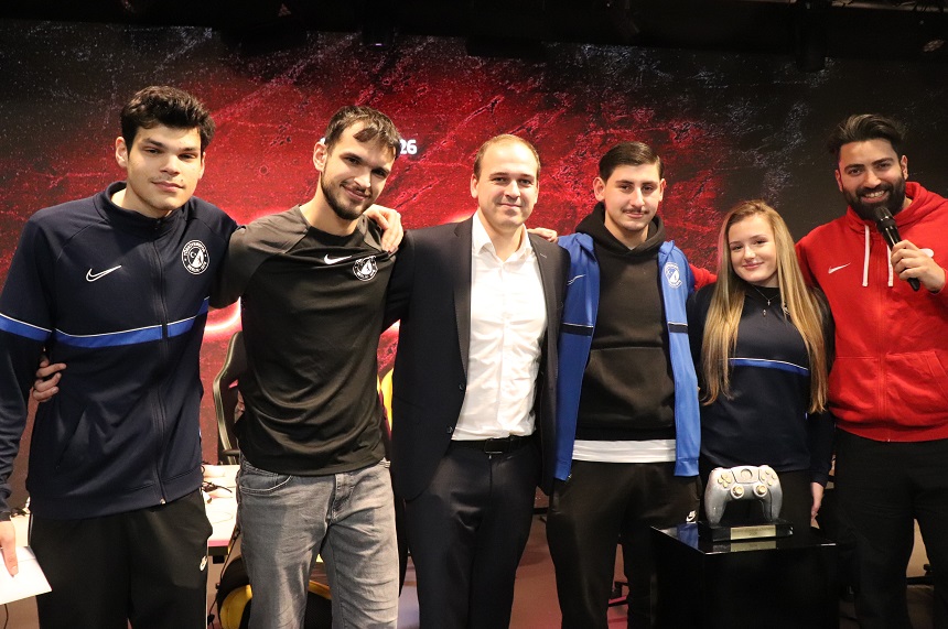 Auf dem Foto stehen sechs Personen eng nebeneinander in einer Reihe auf einer Bühne oder Eventfläche. Sie wirken wie ein Team oder eine Gruppe, die gemeinsam für ein Foto posiert. Einige tragen Sport‑ oder Teamkleidung in Blau oder Schwarz, eine Person trägt einen Anzug, und eine weitere Person trägt eine rote Jacke und hält ein Mikrofon, als würde sie moderieren oder ankündigen. Die Personen stehen vor einem großen Hintergrund mit rot‑schwarzer Grafik, der eine dynamische, sportlich‑technische Atmosphäre vermittelt. Vor der Gruppe steht auf einem kleinen Podest ein großer, auffällig gestalteter Controller, der auf ein Gaming‑ oder E‑Sport‑Event hindeutet. Die Körperhaltung der Gruppe wirkt freundschaftlich und gemeinschaftlich – einige haben die Arme umeinander gelegt, andere stehen nah beieinander, bereit für das gemeinsame Foto.