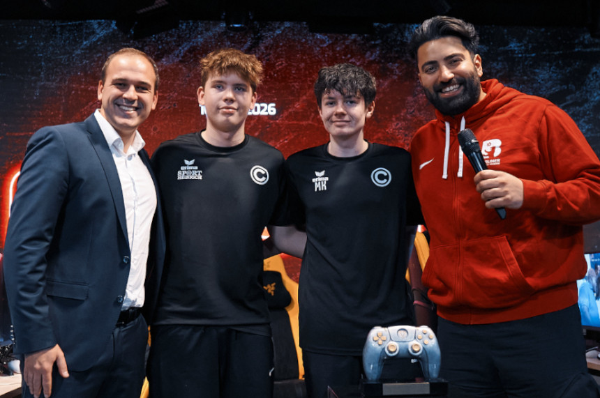 Auf dem Foto stehen vier Personen dicht nebeneinander auf einer Bühne oder vor einer großen Leinwand. Die Hintergrundgrafik zeigt ein dynamisches, dunkel gehaltenes Muster, das an eine E‑Sport‑ oder Gaming‑Veranstaltung erinnert. Drei der Personen tragen schwarze Sport‑ oder Teamshirts mit weißen Logos und Initialen. Sie stehen in einer Reihe und wirken wie Mitglieder desselben E‑Sport‑Teams. Links neben ihnen steht eine Person in einem hellen Anzug, die sich leicht zu ihnen hin orientiert. Rechts daneben hält eine weitere Person in einer roten Jacke ein Mikrofon, als würde sie moderieren oder ein Gespräch führen. Im Vordergrund ist ein großes Gaming‑Pad oder ein spezieller Controller auf einem Ständer zu sehen, was die Szene noch stärker in den Kontext eines Gaming‑Events rückt. Die Körperhaltung aller Beteiligten vermittelt Teamgeist und ein gemeinsames Auftreten bei einer offiziellen Präsentation oder Siegerehrung.