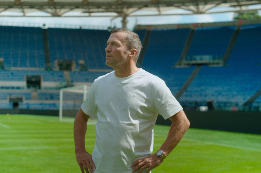 Eine Person im weißen T‑Shirt steht auf einem sonnigen Fußballfeld im Stadion und blickt seitlich über den grünen Rasen. Foto: Tobis