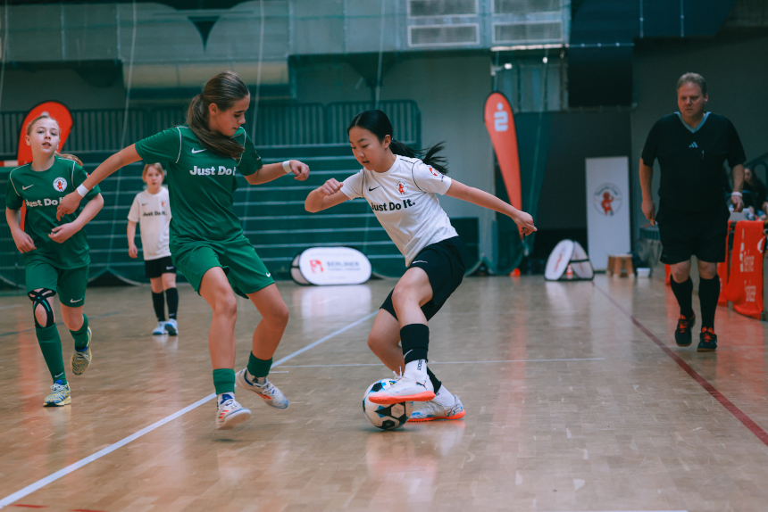 Eine Person in weiß kontrolliert den Ball, während mehrere Personen in grünen Trikots auf dem Hallenboden verteidigen. Foto: Loredana Zafisambondaoky