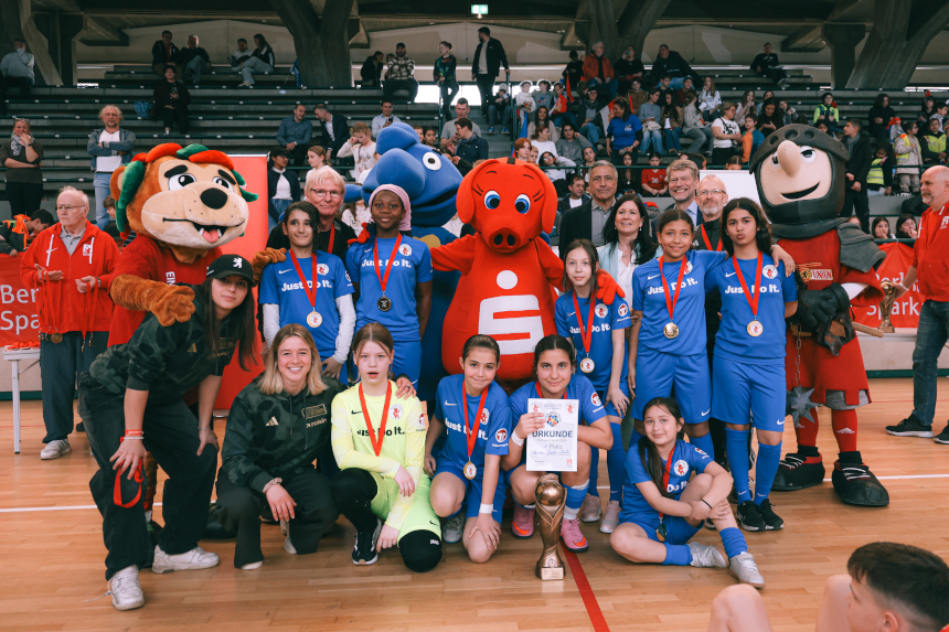 A team in blue jerseys poses with medals and trophy, surrounded by colorful mascots in a crowded sports hall. Photo: Loredana Zafisambondaoky