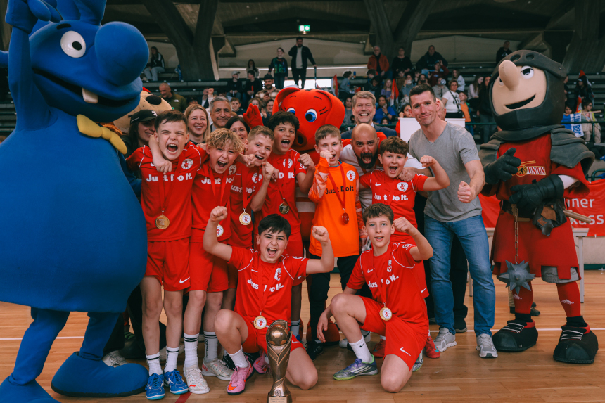 Team in red jerseys poses with medals and trophy, flanked by large colorful mascots in a packed sports hall. Photo: Loredana Zafisambondaoky