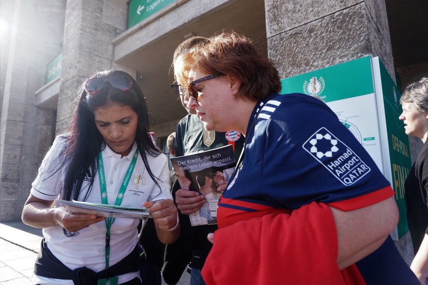 Das Foto zeigt drei Personen, die dicht beieinanderstehen und sich offenbar in ein Gespräch oder eine gemeinsame Aktivität vertieft haben. Die Szene spielt im Freien, vor einem Gebäude mit hellgrauen Steinsäulen und grünen Wegweisern im Hintergrund. Links steht eine Person in einem weißen Shirt mit grünem Akkreditierungsband. Sie hält ein Tablet oder Klemmbrett in beiden Händen und scheint etwas darauf zu zeigen oder zu erklären. Rechts steht eine Person im dunkelblauen Fußballtrikot einer bekannten Vereinsmannschaft. Über ihren Arm hängt ein rotes Kleidungsstück, und sie lehnt sich leicht nach vorn, um besser zu sehen, was gezeigt wird. Zwischen beiden steht eine dritte Person, die ein Heft oder Magazin hochhält. Auf dem Titel ist eine großformatige Überschrift zu lesen und darunter ein Foto mehrerer Menschen. Die Situation wirkt freundlich und kooperativ – als würden die drei Personen Informationen austauschen, etwas überprüfen oder gemeinsam etwas planen, möglicherweise im Kontext eines Fußball-Großereignisses.
