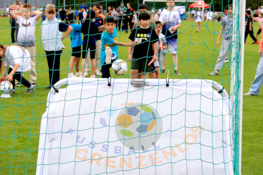 Das Foto zeigt eine lebendige Szene auf einem Fußballfeld während einer öffentlichen Veranstaltung. Im Vordergrund ist das Tornetz eines kleinen Fußballtors zu sehen. Direkt dahinter steht eine weiße Torwand mit einem bunten Logo und der gut lesbaren Aufschrift „FUSSBALL GRENZENLOS“. Ein Fußball befindet sich in der Luft, als wäre er gerade auf das Tor geschossen worden. Hinter dem Tor stehen mehrere Kinder und Jugendliche auf dem Rasen. Einige beobachten das Spiel, andere bewegen sich aktiv und wirken, als würden sie gleich selbst schießen oder spielen wollen. Die Gruppe wirkt vielfältig und bunt gemischt. Im Hintergrund sieht man eine größere Menschenmenge sowie verschiedene Zelte oder Pavillons, die auf ein Sport- oder Gemeinschaftsfest hindeuten. Die Atmosphäre vermittelt Energie, Spaß und ein aktives Miteinander.