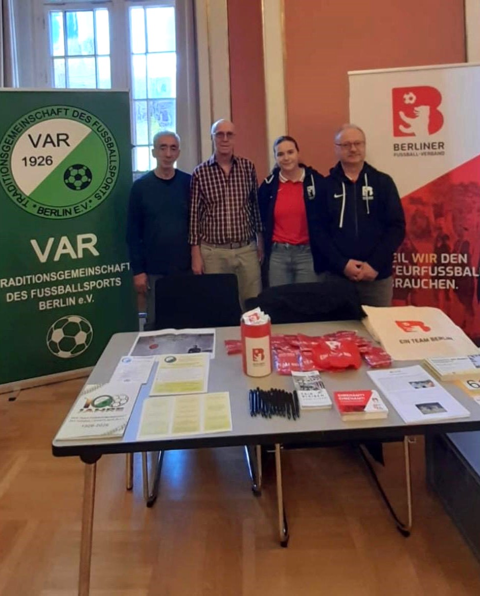 Das Foto zeigt einen Informationsstand in einem Innenraum mit hohen Fenstern und hellem Tageslicht. Im Mittelpunkt stehen vier Personen, die nebeneinander hinter einem Tisch stehen und zur Kamera schauen. Die Personen werden nicht näher beschrieben, sondern sind Teil einer gemeinsamen Darstellung. Vor ihnen steht ein rechteckiger Tisch. Auf dem Tisch liegen verschiedene Informationsmaterialien, darunter Flyer, Broschüren und Ausdrucke. Außerdem sind mehrere schwarze Stifte, rote Stoffbeutel oder Give-aways, ein roter Trinkbecher mit einem Logo sowie eine weiße Stofftasche ausgebreitet. Alles ist ordentlich angeordnet und gut sichtbar. Hinter den Personen stehen zwei große Roll-up-Banner. Der Raum wirkt sachlich und freundlich. Der Boden ist aus Holz, die Wände sind hell- und rosafarben gestrichen. Das Bild vermittelt den Eindruck einer Informations- oder Ehrenamtsbörse, bei der sich Organisationen aus dem Fußballbereich vorstellen. 