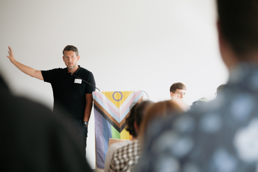 Person at lectern raises arm, speaks in front of seated audience; colorful flag at lectern, bright room, blurred audience in foreground. Photo: LSVD Queer Diversity Association Berlin-Brandenburg 