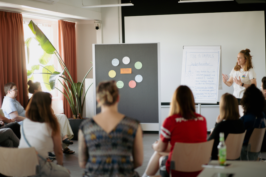 Workshop room: Lecture in front of a seated audience, pinboard with colored cards and flipchart, large plant and window light. Photo: LSVD Queer Diversity Association Berlin-Brandenburg 