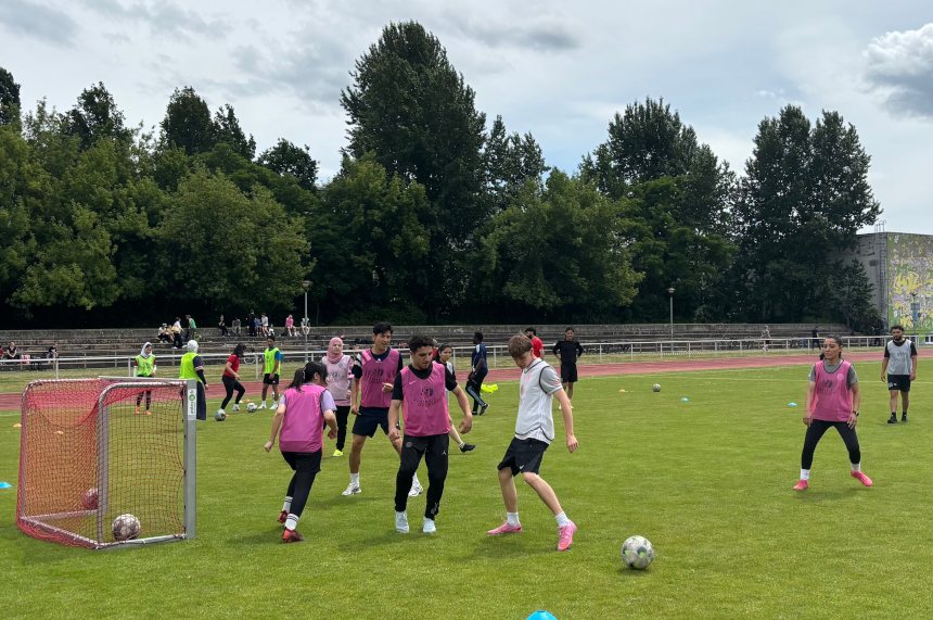 Several people are practicing soccer on a grass field; small goals, lots of balls, pink practice vests, other groups, and bleachers in the background. Photo: BFV