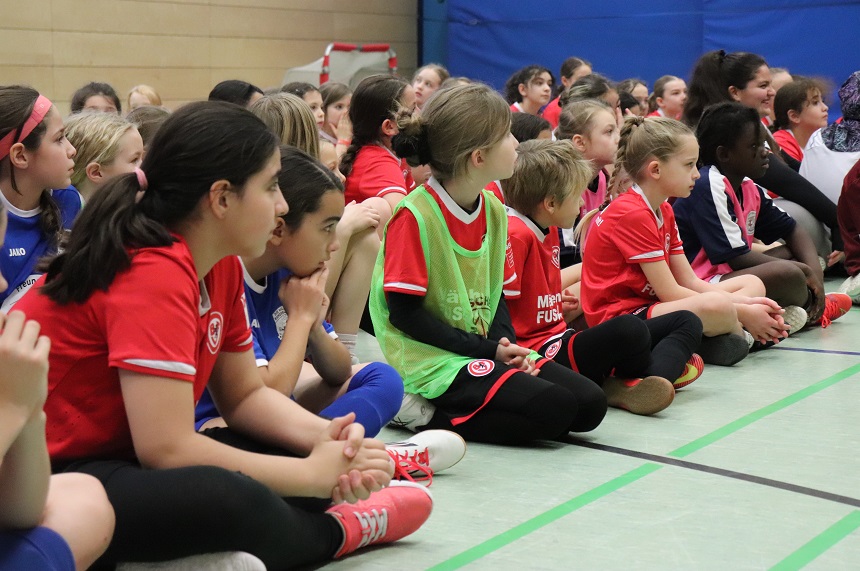 Das Bild zeigt eine Gruppe von Kindern, die in einer Sporthalle auf dem Boden sitzen. Sie tragen überwiegend Sportkleidung, hauptsächlich Trikots in Rot und Blau, kombiniert mit schwarzen oder dunklen Sporthosen. Einige tragen farbige Leibchen, zum Beispiel ein grünes über einem roten Trikot. Die Kinder sitzen in Reihen nebeneinander, die Beine gekreuzt oder angewinkelt, und scheinen aufmerksam in eine Richtung zu schauen, als würden sie einer Ansprache oder Siegerehrung folgen. Der Boden der Halle ist hellgrün mit dunklen Linien, die Spielfeldmarkierungen darstellen. Im Hintergrund sind blaue und beige Wände sowie eine rote Sporttasche zu erkennen. Die Atmosphäre wirkt ruhig und geordnet, typisch für eine Pause oder Abschlussveranstaltung bei einem Turnier.