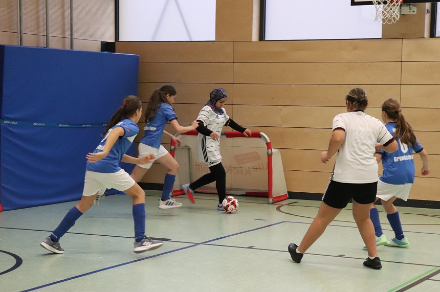 Das Bild zeigt eine Szene in einer Sporthalle während eines Hallenfußballspiels. Fünf Personen sind aktiv am Spiel beteiligt. Sie befinden sich nahe eines kleinen Tores mit rotem Rahmen, das an der Holzverkleidung der Hallenwand steht. Der Boden ist hellgrün mit dunklen Spielfeldlinien. Eine Person in einem weißen Trikot mit schwarzen Streifen, schwarzen langen Hosen und einem gemusterten Kopftuch steht direkt vor dem Tor und kontrolliert den Ball, der rot-weiß gemustert ist. Vier weitere Personen tragen blaue Trikots mit weißen Shorts und blauen Stutzen oder ein weißes Trikot mit schwarzen Shorts. Sie sind in Bewegung und scheinen zu versuchen, den Ball zu erobern. Die Szene wirkt dynamisch, als ob gleich ein Torschuss oder eine Abwehraktion erfolgen könnte. Im Hintergrund sind blaue Matten an der Wand befestigt, darüber Fensterflächen, die Tageslicht hereinlassen. Rechts oben ist ein Basketballkorb zu sehen, was darauf hinweist, dass die Halle für mehrere Sportarten genutzt wird.
