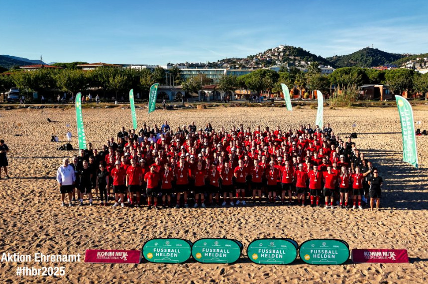 The photo shows a large group of people standing on a wide sandy beach. The group is close together and wearing predominantly red sports shirts with black shorts. Some people on the edge are wearing black or white clothing. In the foreground, several green and pink banners are placed on the sand with the inscriptions "FUSSBALL HEROES" and "KOMM MIT International". At the bottom left of the picture is white text: "Aktion Ehrenamt #fhbr2025". In the background are several green flags with white lettering. Behind them you can see a row of trees, buildings and, further back, a hilly landscape with houses stretching up to the mountain tops. The sky is clear and blue and the sun is shining brightly, indicating warm, sunny weather. The scene gives the impression of a major sporting event or a meeting for soccer volunteers.