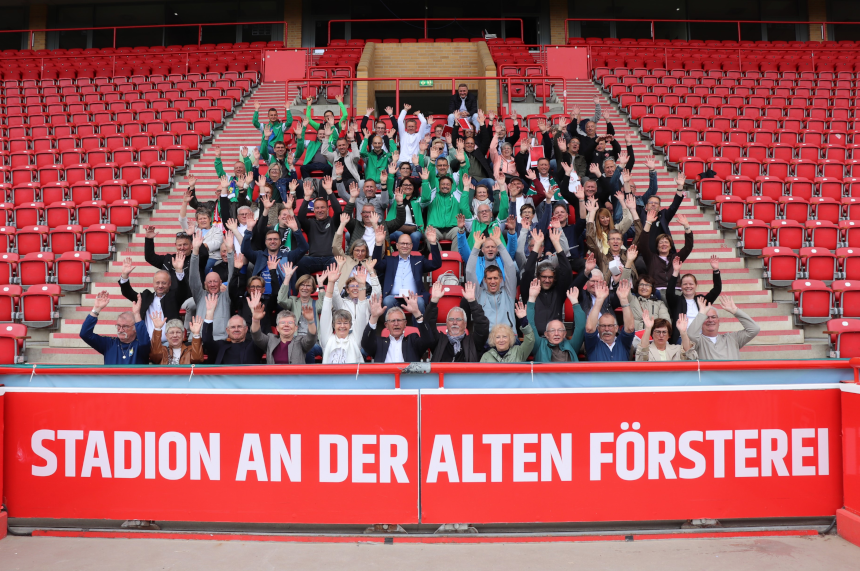 Das Foto zeigt eine große Gruppe von Menschen auf den Tribünen eines Fußballstadions. Die Personen sitzen und stehen auf roten Sitzreihen, die sich stufenförmig nach oben erstrecken. Viele der Anwesenden heben beide Arme nach oben und zeigen offene Hände. Die Personen tragen unterschiedliche Kleidungsstücke, unter anderem Jacken, Pullover, Hemden und Schals in verschiedenen Farben, darunter Grün, Schwarz, Grau und Blau. Einige Personen tragen Brillen, andere Kopfbedeckungen. Die Gruppe wirkt altersmäßig vielfältig. Im unteren Vordergrund befindet sich eine rote Brüstung mit weißer, gut lesbarer Schrift. Darauf steht: „STADION AN DER ALTEN FÖRSTEREI“. Die Schrift ist groß und kontrastreich gestaltet. Im Hintergrund sind weitere Reihen roter Sitzplätze zu sehen, die überwiegend leer sind, sowie Treppenaufgänge mit hellen Stufen und roten Geländern. Die Architektur des Stadions ist klar strukturiert und symmetrisch. Die Szene ist bei Tageslicht aufgenommen, die Beleuchtung ist gleichmäßig, ohne starke Schatten. Das Bild vermittelt den Eindruck einer gemeinsamen Veranstaltung oder eines Gruppentreffens an einem bekannten öffentlichen Ort. Die Menschen sind gleichberechtigt nebeneinander platziert und gemeinsam Teil der Szene.