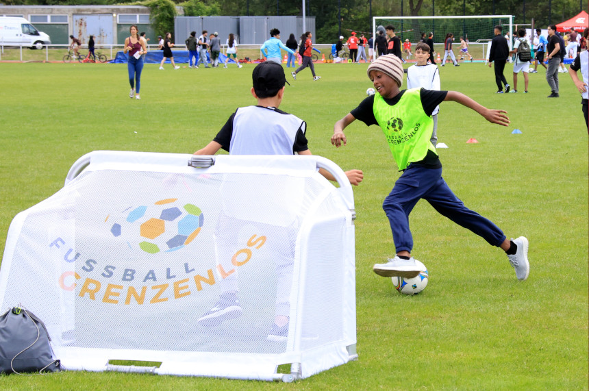 Auf dem Foto ist eine lebhafte Szene auf einem großen Sportplatz zu sehen. Im Vordergrund spielen zwei Kinder Fußball. Eines der Kinder trägt eine helle Trainingsweste und steht direkt vor einem kleinen weißen Popup-Tor, auf dem der Schriftzug „Fussball grenzenlos“ und ein bunter Ball zu sehen sind. Das andere Kind trägt eine neongelbe Trainingsweste und läuft dynamisch auf den Ball zu, als würde es gerade einen Spielzug einleiten oder einen Schuss vorbereiten. Auf der Wiese im Hintergrund befinden sich viele weitere Kinder und Jugendliche, die an unterschiedlichen Bewegungs- und Sportangeboten teilnehmen. Einige laufen, andere spielen oder stehen in Gruppen zusammen. Entlang des Spielfeldrands sind Betreuende und Zuschauende zu erkennen. Die Atmosphäre wirkt fröhlich, offen und von viel Aktivität geprägt. Das Foto vermittelt ein buntes, inklusives Sportfest, bei dem Bewegung, gemeinsames Spielen und Begegnung im Mittelpunkt stehen.