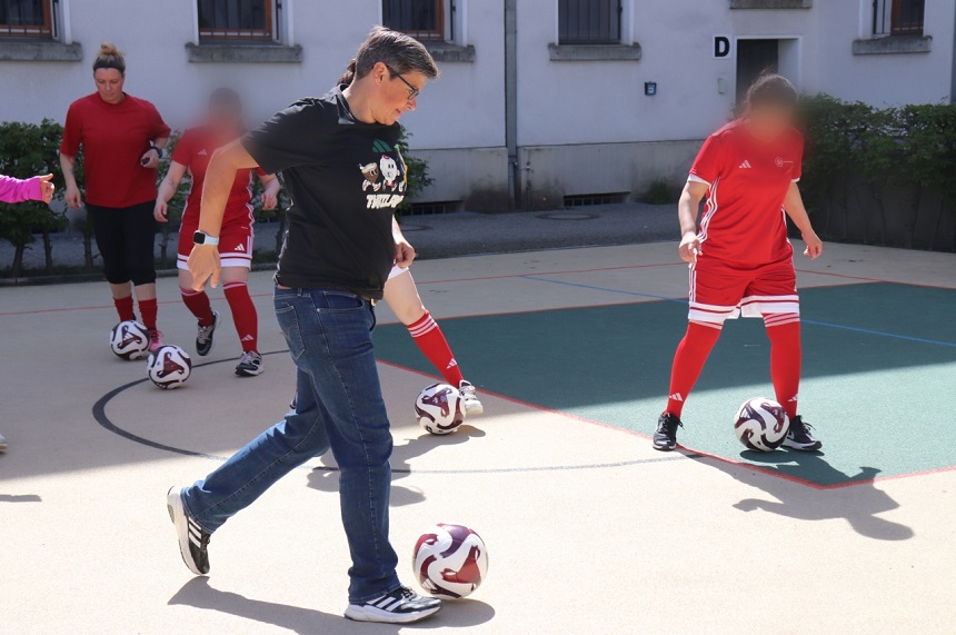Das Foto zeigt eine Gruppe von mehreren Personen auf einer hellen, ebenen Spielfläche im Freien, die mit Linien markiert ist. Jede sichtbare Person hat einen Fußball in der Nähe der Füße; mehrere Bälle liegen gleichmäßig verteilt auf dem Boden. Die Personen bewegen sich gleichzeitig in dieselbe Richtung, was auf eine gemeinsame Übung hindeutet. Im Vordergrund links steht eine Person in Alltagskleidung mit schwarzem Oberteil, Jeans und Sportschuhen. Der Blick und die Körperhaltung sind auf den Fußball am Boden ausgerichtet, ein Fuß ist leicht angehoben, als würde der Ball kontrolliert oder vorwärts bewegt. Im Hintergrund und rechts stehen weitere Personen in roter Sportkleidung mit weißen Streifen, darunter Trikots, Shorts und kniehohe Stutzen. Auch diese Personen führen jeweils einen Ball mit den Füßen. Im Hintergrund ist ein helles Gebäude mit mehreren Fenstern zu sehen, einige davon mit Gitterstäben versehen. Ein Metallzaun und niedrige grüne Büsche grenzen die Spielfläche vom Gebäude ab. An einer Wand ist der Buchstabe „D“ angebracht. Das Licht ist hell und gleichmäßig, was auf sonniges Wetter schließen lässt. Die Gesichter aller Personen sind unscharf dargestellt, sodass keine Identifizierung möglich ist. Die Szene vermittelt den Eindruck eines gemeinsamen Fußballtrainings oder einer angeleiteten Übung, bei der mehrere Personen gleichzeitig und koordiniert mit dem Ball arbeiten.