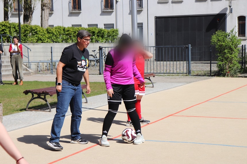 Das Foto zeigt eine sportliche Szene im Freien auf einer ebenen, hellen Spielfläche mit eingezeichneten Linien. Im Vordergrund steht eine Person in einem pinkfarbenen Langarm-Oberteil, schwarzen Shorts und schwarzen Stutzen. Ein Fußball liegt direkt vor den Füßen dieser Person und berührt fast die Schuhspitze. Daneben steht eine weitere Person in Alltagskleidung, bestehend aus einem schwarzen T‑Shirt, blauer Jeans und Sportschuhen, leicht zum Ball hin ausgerichtet. Im Hintergrund befinden sich weitere Personen in roten Sporttrikots und roten Stutzen. Eine Holzbank steht am Rand der Spielfläche. Dahinter ist ein helles Gebäude mit Fenstern zu sehen, das von einem Metallzaun teilweise abgegrenzt wird. Grünflächen, Büsche und ein Baum rahmen den Ort ein. Das Licht ist hell und gleichmäßig, was auf sonniges Wetter hindeutet. Die Gesichter der Personen sind unscharf dargestellt, sodass keine individuellen Merkmale erkennbar sind. Die Szene vermittelt den Eindruck eines Trainings- oder Übungsmoments, bei dem gemeinsam Fußball gespielt oder angeleitet wird.