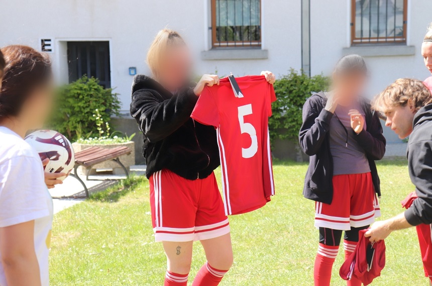 Das Foto zeigt eine Gruppe von mehreren Personen auf einer grünen Rasenfläche im Freien. Die Szene wirkt wie ein sportlicher oder gemeinschaftlicher Moment. Im Mittelpunkt steht eine Person, die ein rotes Sporttrikot mit weißen Streifen an den Seiten und der großen weißen Zahl „5“ auf der Vorderseite hochhält. Mehrere Personen in der Gruppe tragen ähnliche rote Sportkleidung, bestehend aus Trikots, Shorts und kniehohen Stutzen. Links im Bild ist eine Person zu sehen, die einen Fußball in den Händen hält und der Gruppe zugewandt ist. Rechts stehen weitere Personen nah beieinander, eine davon hält ebenfalls ein rotes Kleidungsstück. Alle Gesichter sind unscharf dargestellt, sodass keine Einzelperson identifizierbar ist. Im Hintergrund befindet sich ein helles Gebäude mit Fenstern, teilweise mit Gitterstäben. Davor wachsen Büsche und Pflanzen. Eine Holzbank steht seitlich im Bild. Das Tageslicht ist hell, es gibt klare Schatten, was auf sonniges Wetter hindeutet. Der Gesamteindruck ist eine informelle, gemeinschaftliche Situation rund um sportliche Aktivität oder Teamzugehörigkeit, ohne dass ein konkretes Spielgeschehen zu sehen ist.