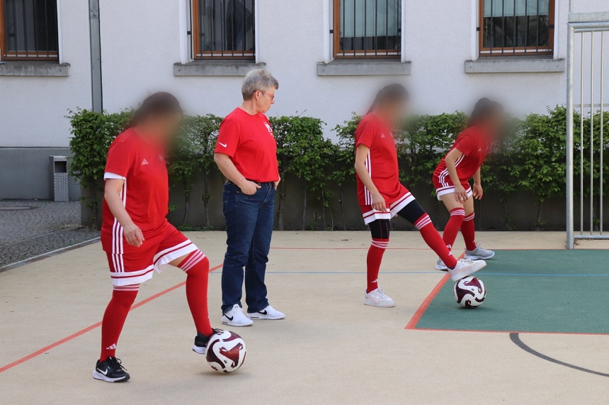 Das Foto zeigt mehrere Personen auf einer ebenen Spielfläche im Freien. Die Fläche ist hell und mit farbigen Linien markiert, was auf ein Sportfeld hindeutet. Alle sichtbaren Personen tragen überwiegend rote Sportkleidung mit weißen Streifen, bestehend aus Trikots, Shorts und kniehohen Stutzen. Eine Person in der Mitte trägt Jeans, ein rotes Oberteil und Sportschuhe und steht zwischen den anderen Personen. Im Vordergrund links steht eine Person mit einem Fußball direkt vor dem Fuß. Rechts davon sind zwei weitere Personen zu sehen, die jeweils einen Fußball kontrollieren, indem ein Fuß auf dem Ball steht oder dicht daneben platziert ist. Die Körperhaltungen wirken konzentriert auf die Bälle gerichtet. Die Gesichter aller Personen sind unscharf dargestellt, sodass keine individuellen Merkmale erkennbar sind. Im Hintergrund befindet sich ein helles Gebäude mit mehreren Fenstern, die mit Gitterstäben versehen sind. Entlang der Gebäudewand wachsen niedrige grüne Büsche. Rechts ist ein Metallzaun zu erkennen, der an die Spielfläche angrenzt. Das Licht ist hell und gleichmäßig, was auf sonniges Wetter schließen lässt. Die Szene vermittelt den Eindruck einer gemeinsamen sportlichen Übung oder Trainingseinheit, bei der mehrere Personen gleichzeitig mit Fußball arbeiten.