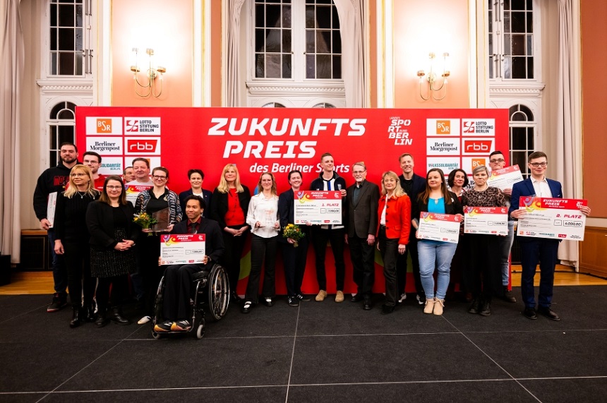 The photo shows a large group of people standing together on a stage in a festive-looking hall. Behind the group is a large red wall with white lettering and logos. The most prominent text reads "Future Prize of Berlin Sports". Several other logos of partners and organizations are visible, including "LSB Berlin", "rbb" and "Berliner Morgenpost". The people stand in two rows next to each other, some holding large symbolic checks, certificates or bouquets of flowers. Among the honorees is also a person in a wheelchair, who stands in the front area and also presents a large cheque. The attire of the people present is predominantly festive or business attire. The room has a prestigious appearance: high, brightly painted walls, large windows with long curtains and classic wall lamps characterize the room. The floor is partially covered with a dark stage platform on which the group stands. Overall, the image conveys the atmosphere of a festive awards ceremony where various people are honored for their achievements or projects in the field of sport.