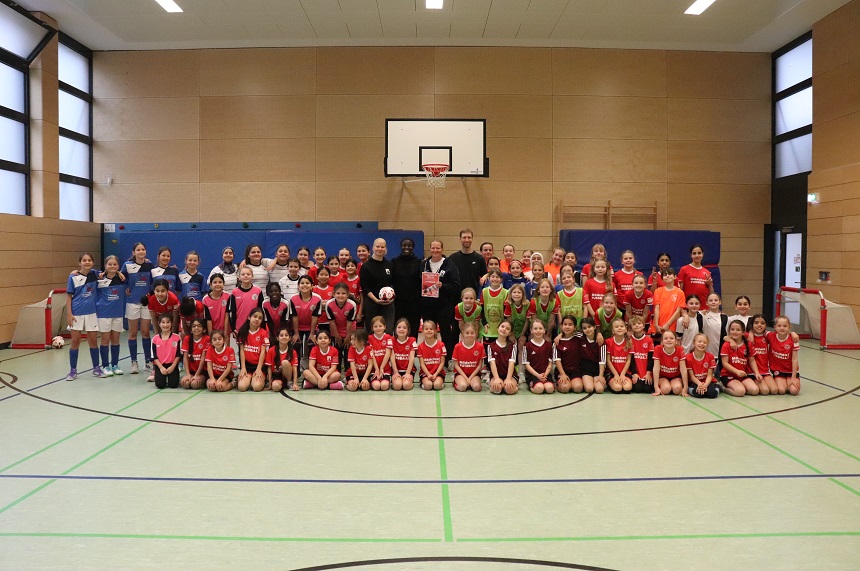 Das Foto zeigt eine große Gruppe von Kindern und Jugendlichen in einer Sporthalle, die für ein gemeinsames Gruppenbild posieren. Die Halle hat helle Wände, große Fenster und einen Basketballkorb an der Rückwand. Der Boden ist grün mit schwarzen und grünen Linien für Spielfelder markiert. Links und rechts am Rand sind kleine Tore zu sehen, was auf ein Fußballtraining oder ein Turnier in der Halle schließen lässt. Die Stimmung wirkt sportlich und gemeinschaftlich, mit einem klaren Fokus auf Teamgeist und Vielfalt.