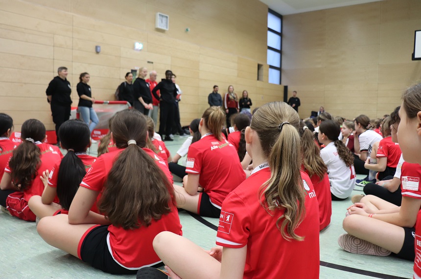 Das Bild zeigt eine Szene in einer Sporthalle mit hellen Holzwänden und großen Fenstern. Im Vordergrund sitzen viele Kinder und Jugendliche auf dem Boden, die meisten tragen rote Sporttrikots mit weißen Aufdrucken. Einige Personen tragen weiße Trikots. Die Gruppe sitzt in lockerer Formation und blickt nach vorne. Im Hintergrund stehen mehrere Erwachsene an der Wand, einige in dunkler Kleidung, andere in Freizeitkleidung. Sie scheinen eine Ansprache oder Präsentation zu halten. Vor ihnen ist ein Tisch oder eine Bank mit roten Taschen oder Materialien darauf. Die Stimmung wirkt konzentriert und gemeinschaftlich. Die Perspektive ist seitlich aufgenommen, sodass die sitzenden Personen von hinten und die stehenden Personen im Hintergrund sichtbar sind. Die Szene vermittelt den Eindruck eines Teamtreffens oder einer Begrüßung vor einem Sportevent.