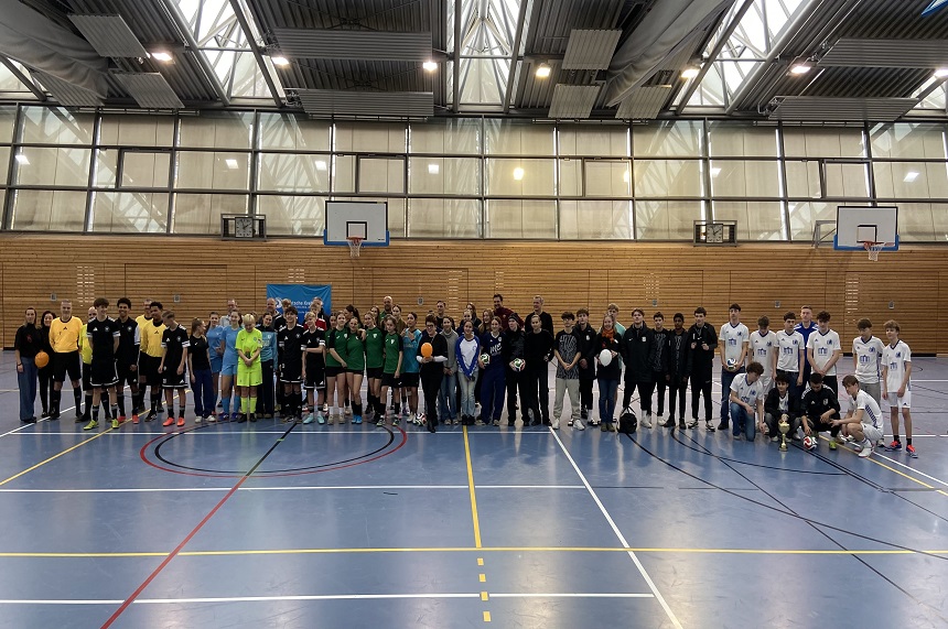 Das Foto zeigt eine große Gruppe von Personen, die in einer Sporthalle aufgereiht steht. Die Sporthalle hat einen hohen Deckenbereich mit vielen Fenstern, durch die Tageslicht hineinfällt. Mehrere Basketballkörbe hängen an den Wänden. Die Personen stehen in einer langen Reihe quer durch die Mitte des Bildes. Sie tragen überwiegend Sportkleidung in unterschiedlichen Farben, unter anderem Schwarz, Grün, Blau und Weiß. Einige tragen Trikots, andere Trainingskleidung. Mehrere Personen halten Fußbälle oder Handbälle in den Händen. Die Gruppe wirkt vielfältig: Es sind Menschen unterschiedlicher Körpergrößen und Altersstufen zu sehen. Im Hintergrund ist eine Holzwand zu erkennen, wie sie typischerweise in Sporthallen vorkommt. Auf der linken Seite steht ein blaues Banner, dessen Text im Foto nicht lesbar ist. Der Boden ist ein glatter Hallenboden in Blau mit farbigen Spielfeldmarkierungen in Gelb, Rot und Schwarz.