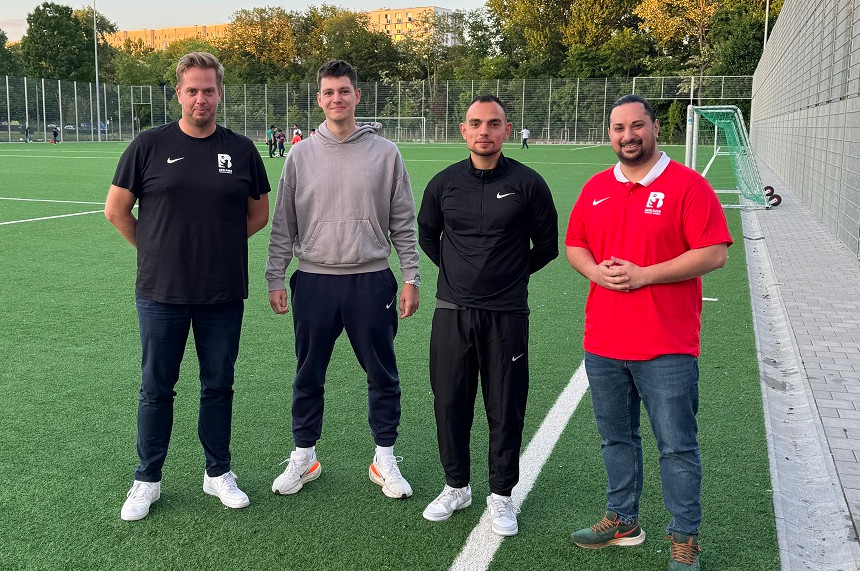 Das Foto zeigt vier Personen, die nebeneinander auf einem Fußballfeld stehen. Sie blicken direkt in die Kamera und wirken präsent und aufmerksam. Die Personen unterscheiden sich in Kleidung, Körperbau und Erscheinung, was auf eine vielfältige Gruppe hinweist.  Die erste Person von links trägt ein schwarzes T-Shirt mit einem weißen Logo und eine dunkle Hose. Die zweite Person hat einen grauen Kapuzenpullover und eine dunkle Hose an. Die dritte Person ist ganz in Schwarz gekleidet – mit einem schwarzen Oberteil und einer schwarzen Hose. Die vierte Person trägt ein rotes T-Shirt mit einem weißen Logo und eine blaue Jeanshose. Im Hintergrund sind weitere Menschen zu sehen, die sich auf dem Fußballfeld bewegen oder spielen. Das Feld ist von einem hohen Zaun umgeben, und dahinter stehen einige Bäume. Die Szene spielt sich im Freien bei Tageslicht ab.
