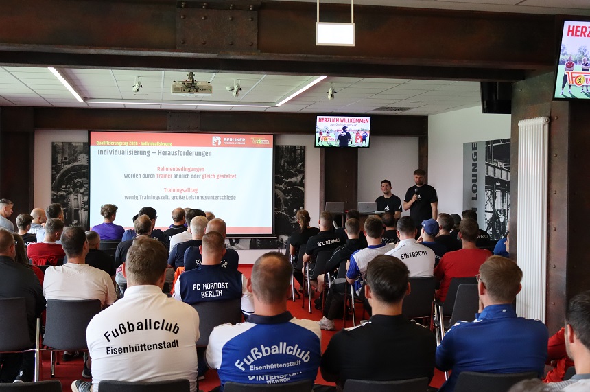 The photo shows an event hall where a presentation is being given to an audience. In the foreground, several rows of chairs are visible, with numerous people sitting on them and looking forward toward a screen. Most of the people are wearing athletic clothing, often with club names or logos on the back, such as “Eisenhüttenstadt Soccer Club” or “FC Nordost Berlin.” In the center of the image, two people are standing in front of the presentation area. One person is standing at the lectern or table with a laptop, while the other person is standing next to them, holding a microphone. Both appear to be speakers or moderators for the event. In the background, a large projection screen displays a presentation slide. Above the screen is a banner with the text “WELCOME.” Logos and a reference to “Qualification 2026 – Individualization” are also visible. A small screen also displays the welcome message and graphic elements. The room has a modern design, with a light-colored ceiling, lighting fixtures, and exposed support beams made of dark material. On the right side, an area labeled “LOUNGE” is visible, as well as a radiator on the wall. The scene conveys a training session or seminar in a sports context, where participants from various clubs are listening to a presentation.