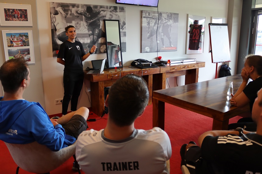 The photo shows a small seminar or meeting room with a sports theme. In the foreground, several people are seated on chairs and facing the front of the room. One person in the foreground is wearing a light-colored T-shirt with the clearly legible word “TRAINER” on the back. Other seated people are wearing athletic clothing such as track jackets or T-shirts. In the center of the image, a person is standing in front of the group and giving a presentation. The standing person is wearing a dark sports outfit and is pointing with one hand toward the wall or screen, while the other hand is in a relaxed position. Next to the person is a table with a laptop and other equipment, including a phone and smaller devices. On the wall behind the standing person hang several large-format pictures with soccer motifs. The pictures show game scenes and are predominantly in shades of gray. Between the pictures is a mirror in which parts of the room are reflected. On the right wall, a framed jersey with the number “28” is visible. On the right side of the room stands a flipchart with blank paper. Next to it is a larger wooden table on which, among other things, a clear bottle is placed. The furnishings appear modern and functional, with light-colored walls, wooden furniture, and red carpet. Overall, the scene conveys a small training or learning situation in a sports setting, where knowledge is imparted in an informal group atmosphere.