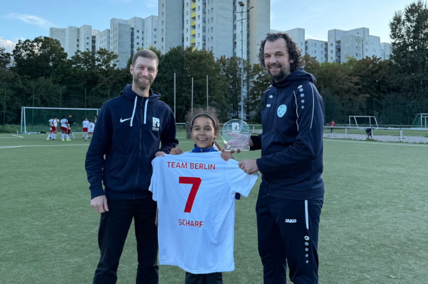 The picture shows three people on a soccer pitch. The person in the middle is holding a white jersey with red lettering up to the camera. The top of the jersey reads "TEAM BERLIN" and below it the large red number "7" and the name "SCHARF". The two people on the left and right stand close to the middle person and touch the jersey. The person on the right is also holding a transparent trophy. All three are wearing sportswear, two of them dark tracksuits with logos. In the background, other players in white jerseys with red shorts can be seen standing in a group. The soccer pitch is surrounded by a high fence, with several tall residential buildings and trees behind it. The sky is clear and there is daylight.