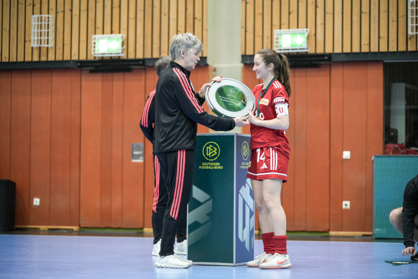 Eine Person in roter Sportkleidung erhält in einer Halle einen Pokalteller von einer Person in schwarzer Trainingskleidung. Foto: 1. FC Union Berlin