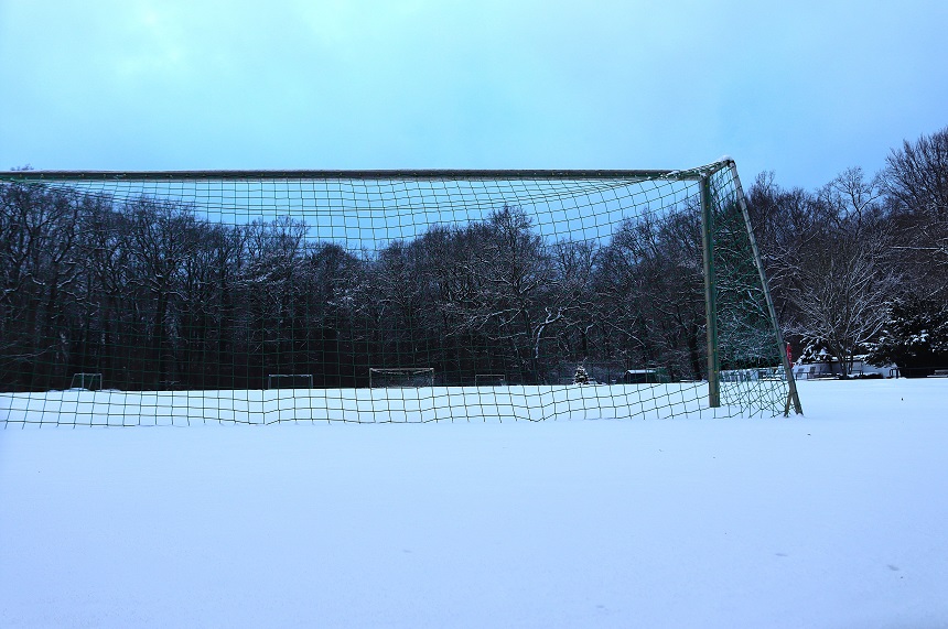The photo shows a single soccer goal in the foreground, taken from a low perspective just above the ground. The entire ground is covered by a thick, untouched layer of snow, forming an even white surface. The soccer goal consists of a metal frame with a green net that is slightly sagging. The snow also lies at the bottom of the net and partially touches the posts. In the background is a dense row of trees whose branches are also covered in a thin layer of snow. The trees appear bare in winter. The sky above is light blue to gray and appears cloudy, giving the scene a calm and cold atmosphere. Overall, the photo gives the impression of a completely snow-covered, quiet sports field in winter, with no visible people or animals.