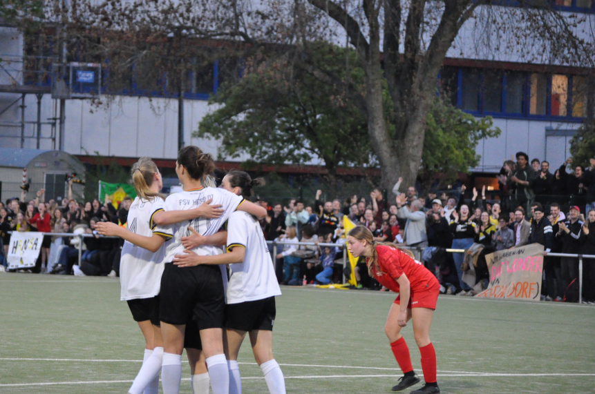 Mehrere Fußballspieler*innen in weißen Trikots umarmen sich auf dem Spielfeld, im Hintergrund jubeln Zuschauende, daneben steht eine Person im roten Trikot. Foto: BFV