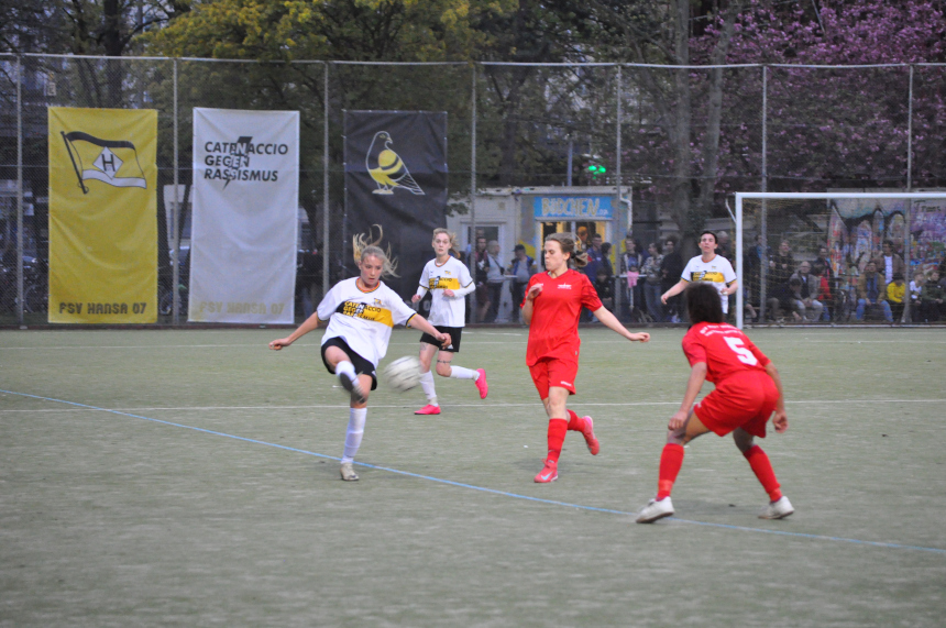Mehrere Fußballspielende kämpfen um den Ball auf dem Spielfeld; weiße und rote Trikots, dahinter Banner und Zuschauende am Zaun. Foto: BFV