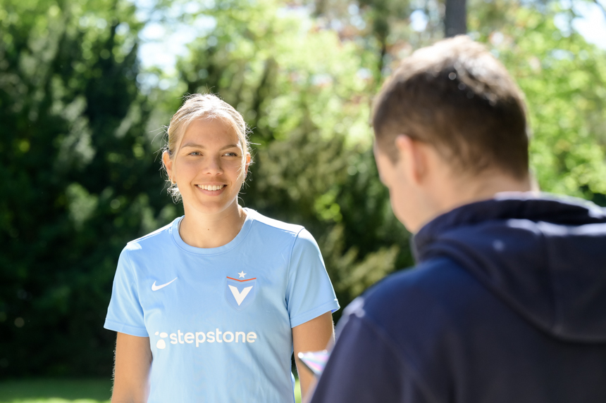 Das Foto zeigt zwei Personen, die sich im Freien unterhalten. Die Person auf der linken Seite trägt ein hellblaues T-Shirt. Auf der rechten Brustseite des T-Shirts befindet sich ein weißes Nike-Logo, auf der linken Seite ein Logo mit einem roten Stern und einem blauen „V“ sowie darunter der Schriftzug „stepstone“. Die Person auf der rechten Seite ist von hinten zu sehen und trägt eine dunkelblaue Jacke oder einen Pullover. Im Hintergrund sind grüne Bäume und Sträucher zu erkennen. Die Szene spielt sich bei sonnigem Wetter ab.