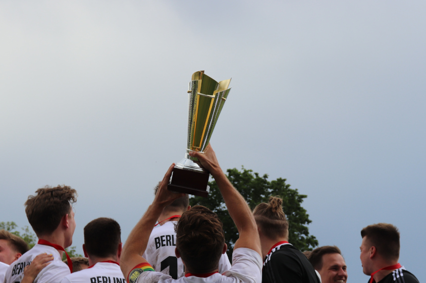 Mehrere Personen in weißen Trikots heben gemeinsam einen goldenen Pokal in die Höhe, Medaillen hängen um ihre Hälse vor hellem Himmel. Foto: BFV