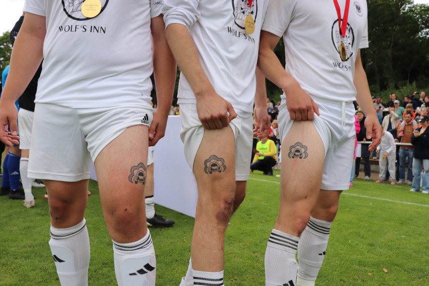 Drei Personen in weißen Trikots zeigen ähnliche Tattoos am Oberschenkel, Medaillen hängen um den Hals, im Hintergrund stehen Zuschauende am Spielfeldrand. Foto: BFV