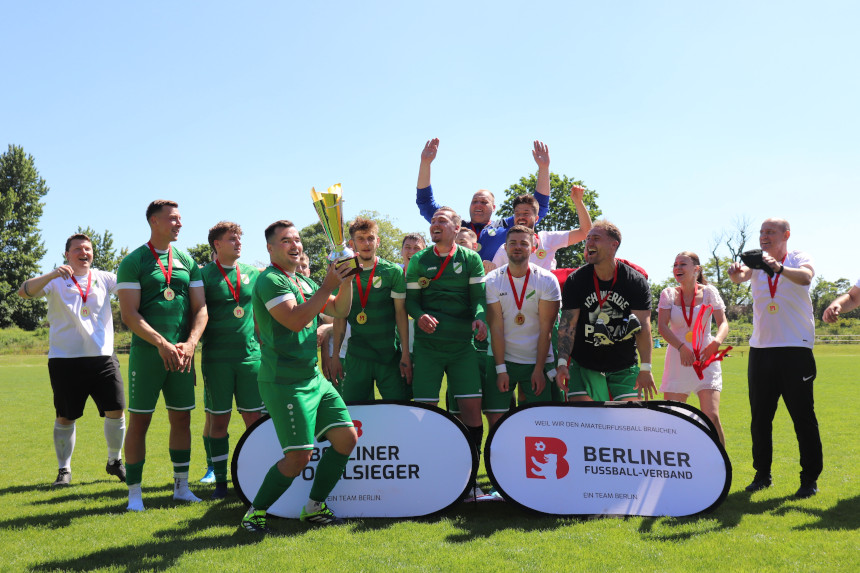 Ein Team in grünen Trikots mit Medaillen steht auf dem Rasen, eine Person hält einen Pokal, im Hintergrund Banner des Berliner Fußball-Verbands. Foto: BFV
