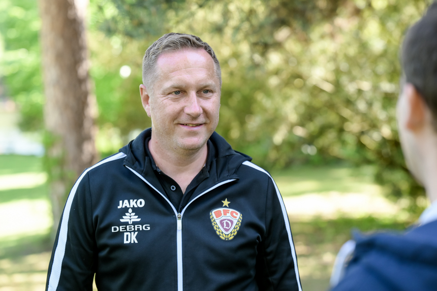 Das Foto zeigt zwei Personen im Freien, vermutlich in einem Park oder einem bewaldeten Gebiet. Im Vordergrund steht eine Person, die der Kamera zugewandt ist. Die Person trägt eine schwarze Jacke mit weißen Streifen an den Schultern und Ärmeln. Auf der Jacke befinden sich mehrere Logos und Schriftzüge, darunter „JAKO“, „DEBRG“, „DK“ sowie ein Wappen mit der Aufschrift „DFC“. Die zweite Person steht mit dem Rücken zur Kamera und ist nur teilweise sichtbar. Im Hintergrund sind Bäume und viel Grün zu sehen, was auf eine natürliche Umgebung hinweist.