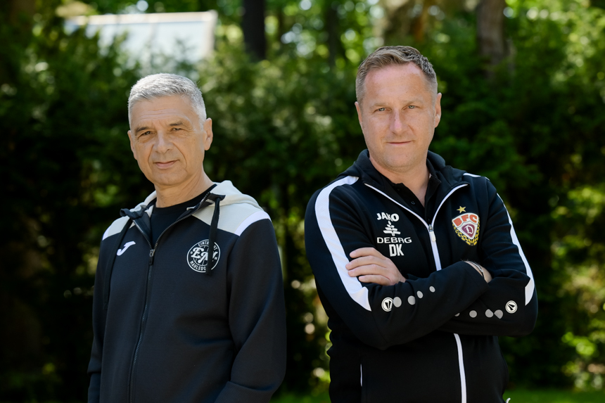 Auf dem Foto sind zwei Personen zu sehen, die nebeneinander im Freien stehen. Beide tragen Sportkleidung. Die Person auf der linken Seite trägt eine schwarze Jacke mit einem weißen Reißverschluss. Auf der Brust der Jacke befindet sich ein Logo. Die Person auf der rechten Seite trägt ebenfalls eine schwarze Trainingsjacke, die weiße Streifen an den Ärmeln hat. Auf ihrer Jacke sind mehrere Logos zu erkennen, darunter ein Wappen und die Buchstaben „DK“. Der Hintergrund zeigt grüne Bäume und Sträucher, was auf eine natürliche Umgebung hinweist.
