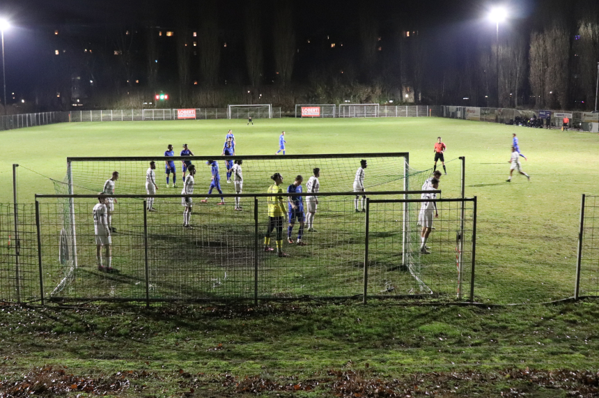 Das Foto zeigt eine nächtliche Fußballszene auf einem beleuchteten Spielfeld. Im Vordergrund ist ein Tor mit Netz zu sehen, um das sich mehrere Spieler gruppiert haben. Einige Spieler tragen weiße Trikots, andere blaue Trikots. Direkt vor dem Tor steht ein Spieler in gelbem Trikot, vermutlich der Torwart. Die Spieler wirken konzentriert, als ob sie sich auf eine Standardsituation wie eine Ecke oder einen Freistoß vorbereiten. Der Rasen ist grün, aber leicht uneben und zeigt Spuren vom Spiel. Im Hintergrund sind weitere Spieler verteilt, ein Schiedsrichter in rotem Trikot ist ebenfalls sichtbar. Hinter dem Spielfeld stehen hohe, kahle Bäume, und am oberen Bildrand leuchten einige Fenster von Gebäuden. Die Szene wird von starken Flutlichtstrahlern erhellt, die deutliche Schatten auf dem Boden werfen.