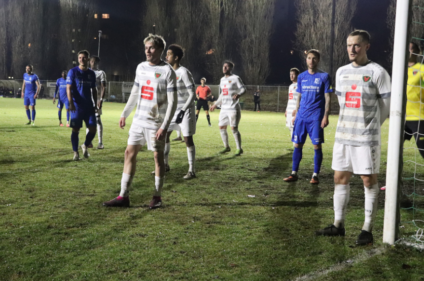 Das Bild zeigt eine Szene auf einem Fußballfeld bei einem Abendspiel unter Flutlicht. Mehrere Spieler sind zu sehen, die sich nahe am Tor positioniert haben. Die Spieler tragen zwei unterschiedliche Trikotsätze: eine Gruppe in weißen Trikots mit grauen Querstreifen und einem großen roten „S“-Logo auf der Brust, die andere Gruppe in blauen Trikots mit weißer Schrift. Ein Torpfosten ist am rechten Bildrand sichtbar, daneben steht ein Spieler in weißem Trikot direkt vor dem Netz. Hinter ihm ist ein Torwart in gelbem Trikot zu erkennen. Im Hintergrund sind hohe, kahle Bäume und einige Lichtpunkte von Gebäuden oder Straßenlaternen sichtbar. Der Rasen ist grün, aber leicht uneben und zeigt Spuren vom Spiel. In der Mitte des Bildes steht ein Schiedsrichter in einem roten Trikot, der die Szene beobachtet. Die Beleuchtung ist hell, aber typisch für ein Flutlichtspiel, mit starken Schatten auf dem Boden.