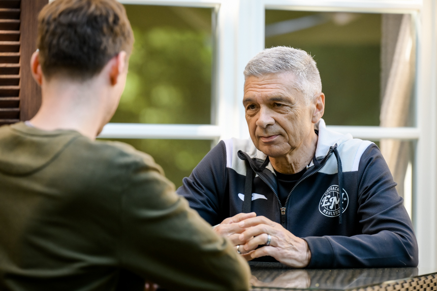 Zwei Personen sitzen an einem Tisch und unterhalten sich. Die Person rechts trägt eine dunkle Jacke mit weißen Akzenten und Logo von Eintracht Mahlsdorf. Es handelt sich um Trainerlegende Karsten Heine. Die Person links, mit dem Rücken zur Kamera, hat kurze braune Haare und trägt ein grünes Oberteil. Diese Person interviewt Mahlsdorf-Coach Karsten Heine. Sie befinden sich in einem Innenraum mit einem Fenster, durch das grüne Pflanzen zu sehen sind. Foto: Sandra Ritschel