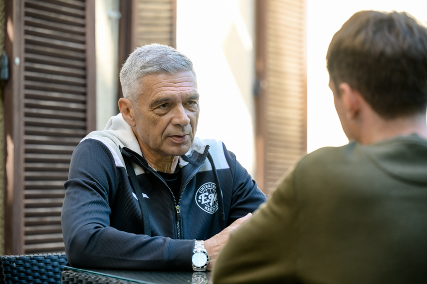 Zwei Personen sitzen an einem Tisch und unterhalten sich. Die Person rechts trägt eine dunkle Jacke mit weißen Akzenten und Logo von Eintracht Mahlsdorf. Es handelt sich um Trainerlegende Karsten Heine. Die Person links, mit dem Rücken zur Kamera, hat kurze braune Haare und trägt ein grünes Oberteil. Diese Person interviewt Mahlsdorf-Coach Karsten Heine. Sie befinden sich in einem Innenraum mit einem Fenster, durch das grüne Pflanzen zu sehen sind. Foto: Sandra Ritschel