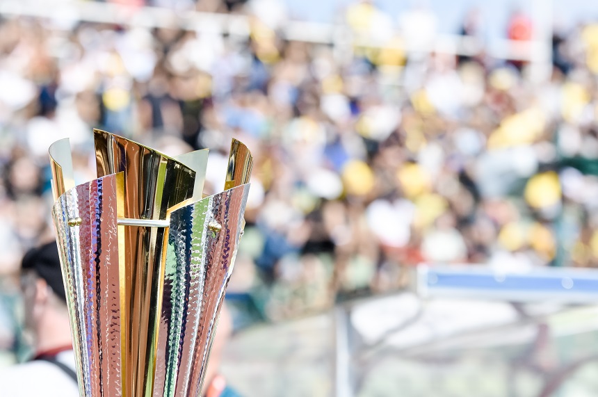 Im Vordergrund des Fotos steht eine Pokaltrophäe aus Metall. Die Trophäe ist hoch und schlank geformt und besteht aus mehreren leicht gebogenen Elementen, die nach oben hin geöffnet sind. Die Oberfläche wirkt glänzend und reflektierend, mit gold- und silberfarbenen Tönen, die je nach Lichteinfall schimmern. Das Material scheint strukturiert zu sein und wirft kleine Lichtreflexe. Im Hintergrund ist unscharf eine große Menschenmenge zu erkennen, vermutlich auf einer Tribüne oder in einem Stadion. Die vielen Farben – unter anderem Gelb, Grün und Weiß – deuten auf Kleidung, Fahnen oder Sitzplätze hin. Einzelne Personen sind nicht erkennbar, da der Fokus des Bildes klar auf der Trophäe liegt. Die Szene findet offenbar im Freien statt, bei Tageslicht. Das Bild vermittelt den Eindruck eines sportlichen Wettbewerbs oder einer Siegerehrung, bei der die Trophäe stellvertretend für Erfolg, Leistung oder einen gewonnenen Titel steht.