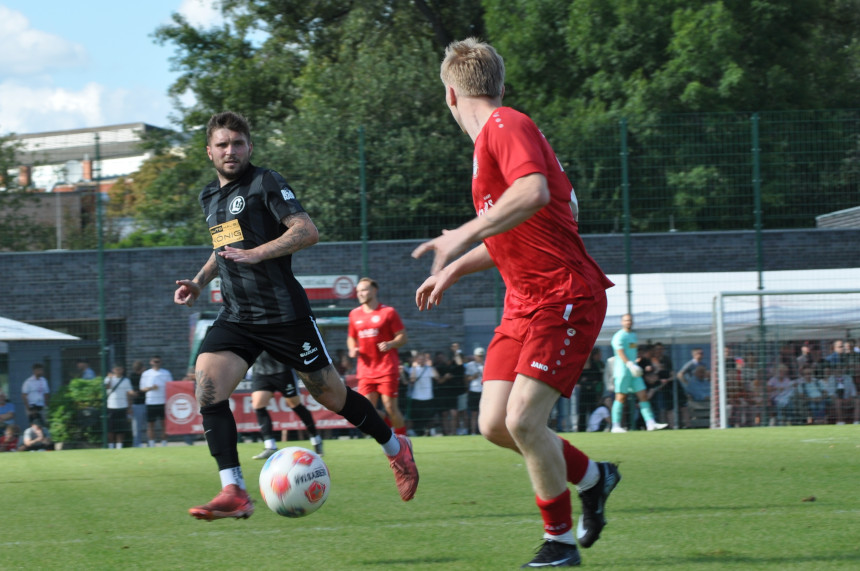 Das Foto zeigt eine Szene von einem Fußballspiel auf einem grünen Rasenplatz. Im Vordergrund befinden sich zwei männlich gelesene Spieler: Einer trägt ein schwarzes Trikot und hat den Ball am Fuß, der andere trägt ein rotes Trikot und läuft hinter ihm her. Der Spieler im schwarzen Trikot scheint sich in Richtung des gegnerischen Tores zu bewegen. Im Hintergrund sind weitere Spieler auf dem Spielfeld verteilt, sowie Zuschauer am Spielfeldrand. Ein Fußballtor ist ebenfalls sichtbar. Die Umgebung des Spielfelds ist von Bäumen und Gebäuden umgeben, was auf eine städtische oder schulische Anlage hindeuten könnte. Das Wetter wirkt freundlich und klar.